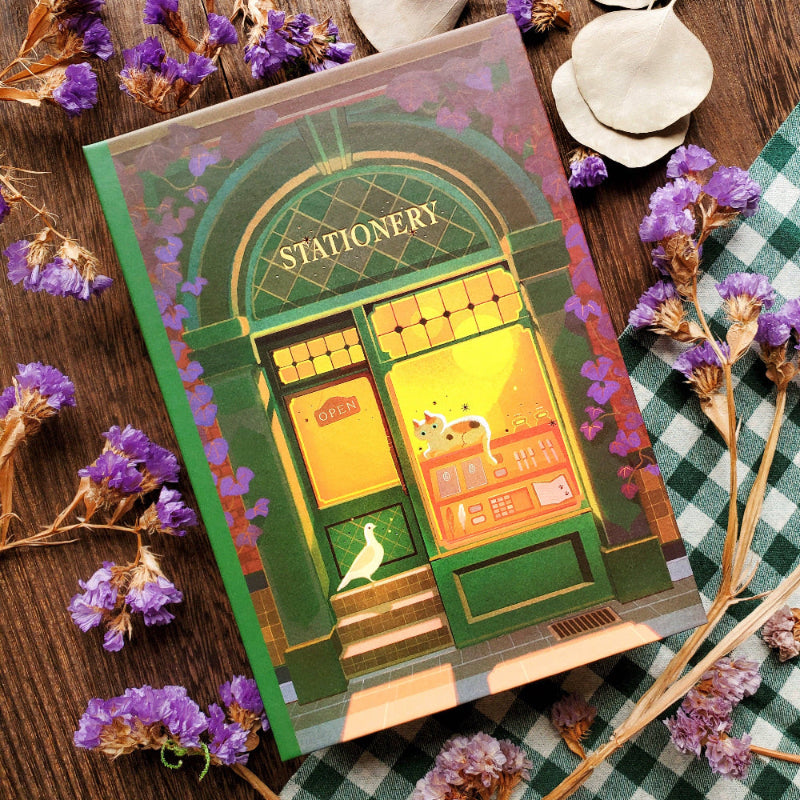 Stationery-themed notebook with a cat illustration on a wooden surface with purple flowers.