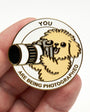 A hand holding a round enamel pin featuring a small fluffy dog holding a camera, with the text 'YOU ARE BEING PHOTOGRAPHED' on a white background.