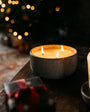 Two lit candles in textured holders on a wooden surface with blurred lights in the background