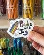 Hand holding a 'Pride and Joy' sticker with colorful design in front of stationery items.