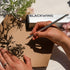 Person holding a Blackwing pencil with a box and flowers in the background