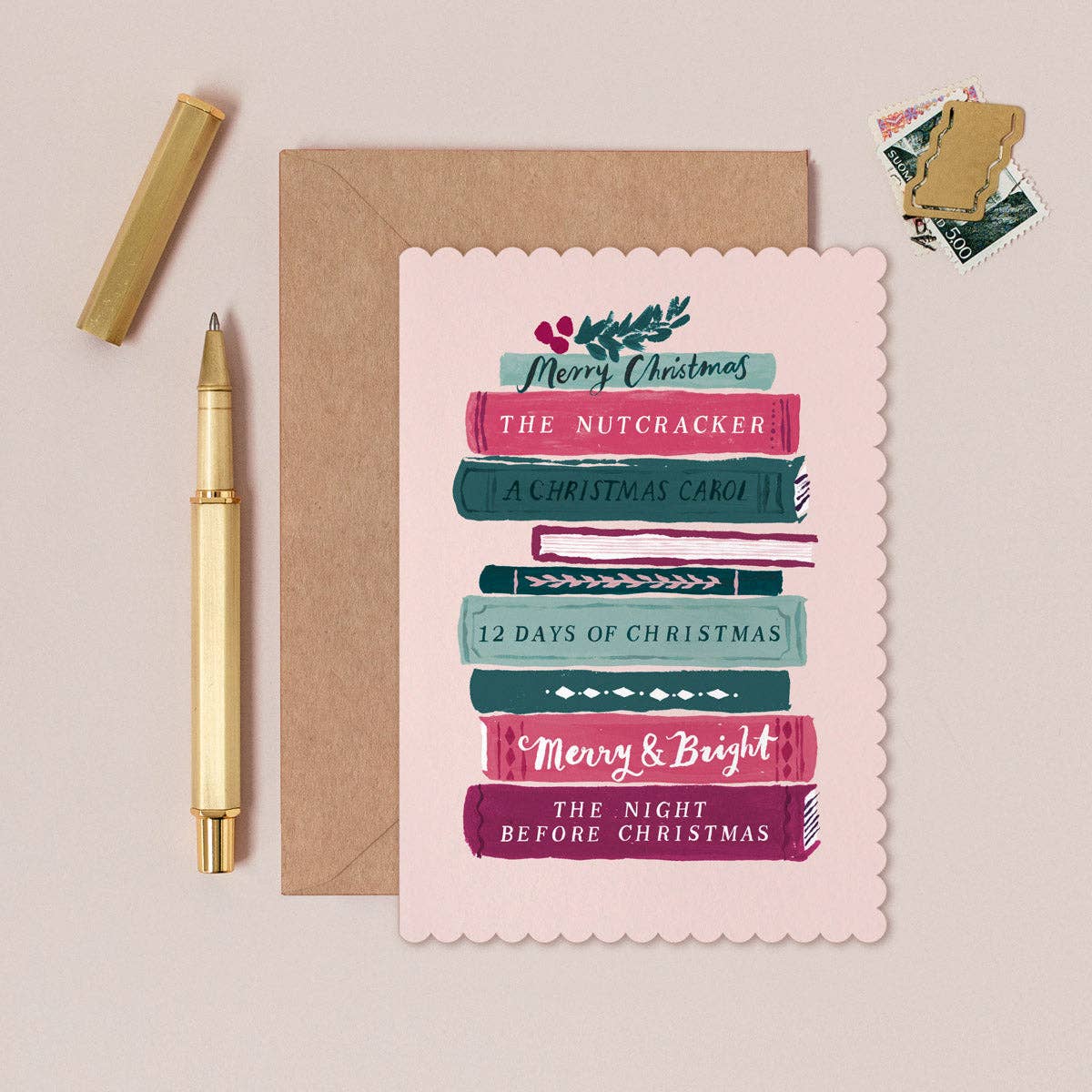 A scalloped-edge Christmas greeting card with stacked book illustrations and holiday phrases, shown with a brown envelope on a beige background. A gold pen, a gold cap, and a small stack of vintage-style postage stamps are arranged around it.