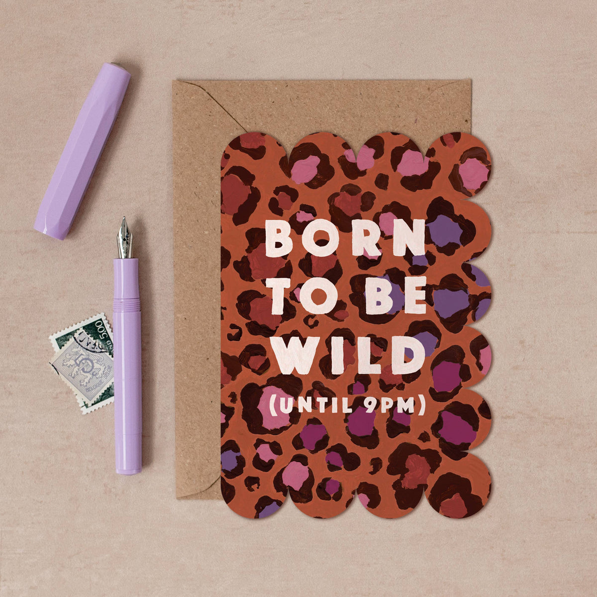 A scalloped greeting card with a brown, pink, and purple leopard print background and the text 'BORN TO BE WILD (UNTIL 9PM)' in white. The card is displayed on a kraft paper envelope with a lavender pen, a lavender cap, and a vintage postage stamp on a beige textured background.