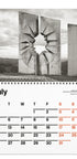 A spiral-bound wall calendar page showing a black-and-white photograph of a large concrete monument with a cracked opening, above a July calendar grid with black and orange dates.