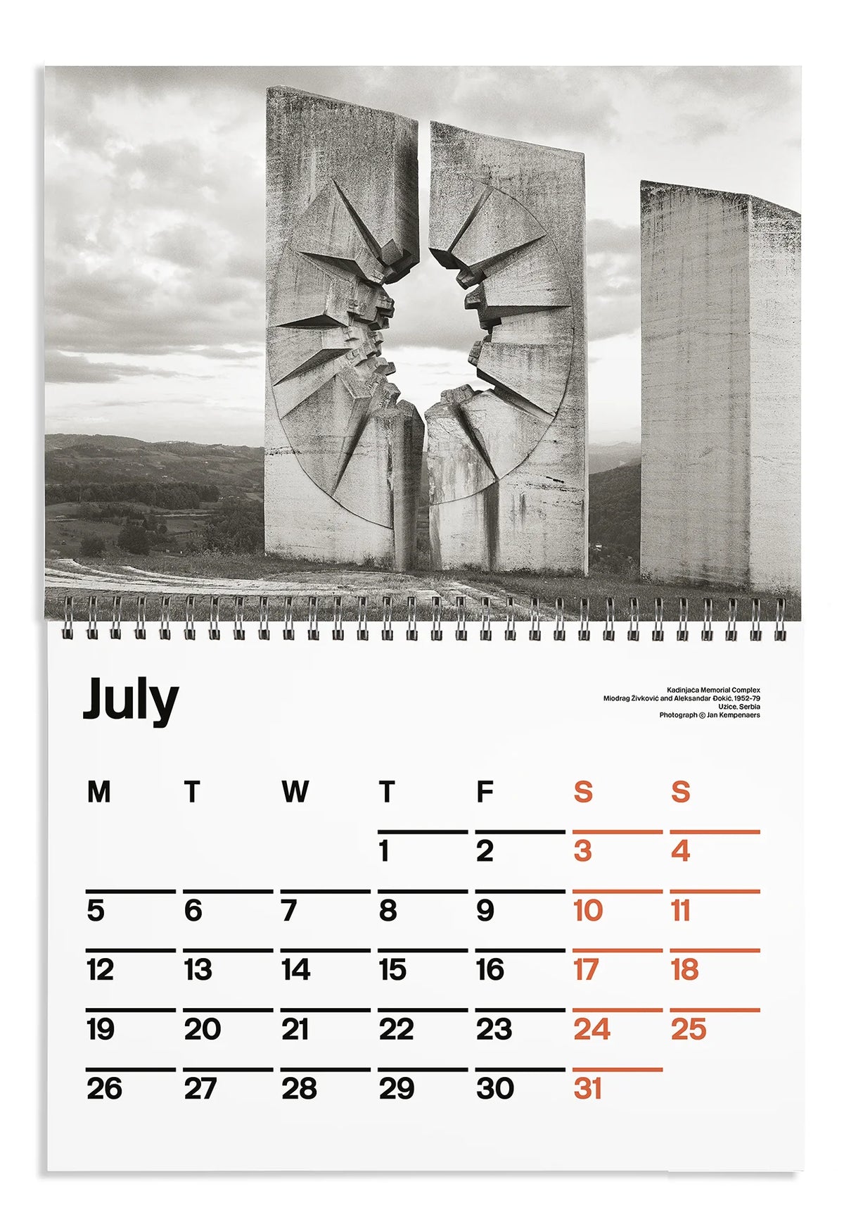A spiral-bound wall calendar page showing a black-and-white photograph of a large concrete monument with a cracked opening, above a July calendar grid with black and orange dates.