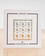 A square white card featuring a 'Camera Shutter Speed Guide' with black border lines and a central gold-bordered panel showing shutter speeds as icons. The card is displayed upright on a light wooden surface against a white background.