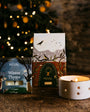 Two candles with decorative boxes on a wooden surface in front of a Christmas tree.