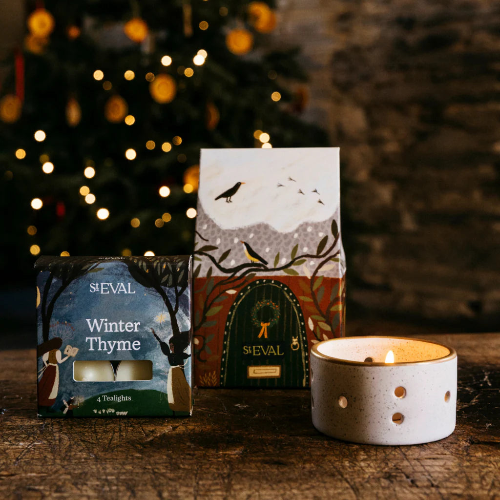Two candles with decorative boxes on a wooden surface in front of a Christmas tree.
