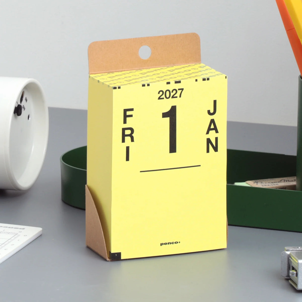 A yellow desk calendar page standing upright on a gray tabletop, with brown cardboard hanger tabs and black printed text showing 2027, Friday, 1, and Jan. Surrounding objects include a white cup, a green container, and office supplies.