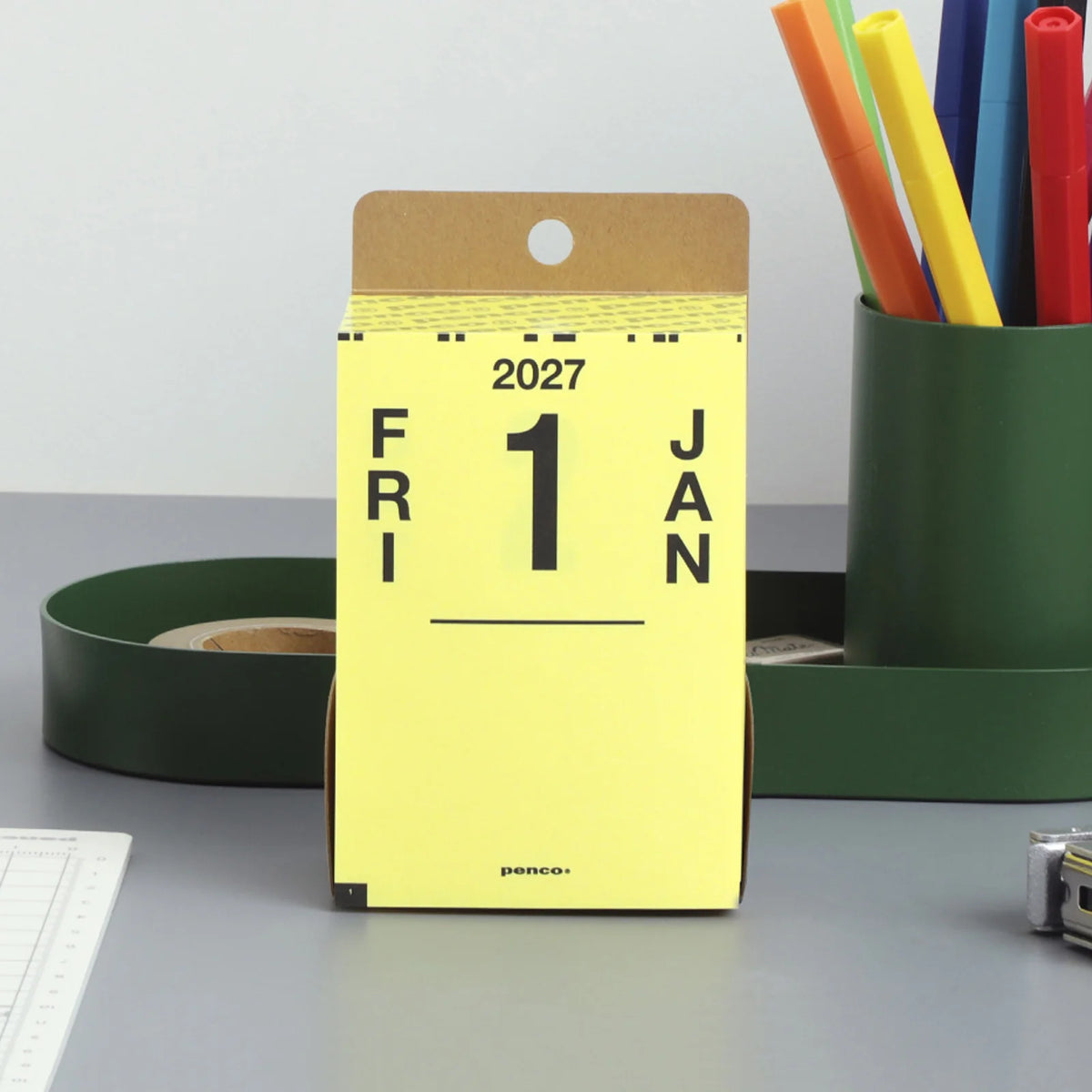 A yellow Penco 2027 desk calendar showing the 1st of January, standing on a gray desk with office supplies in the background.