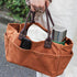 Brown tote bag with handles held by a person, containing a thermos and phone case.