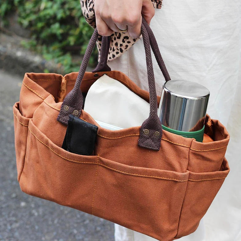 Brown tote bag with handles held by a person, containing a thermos and phone case.