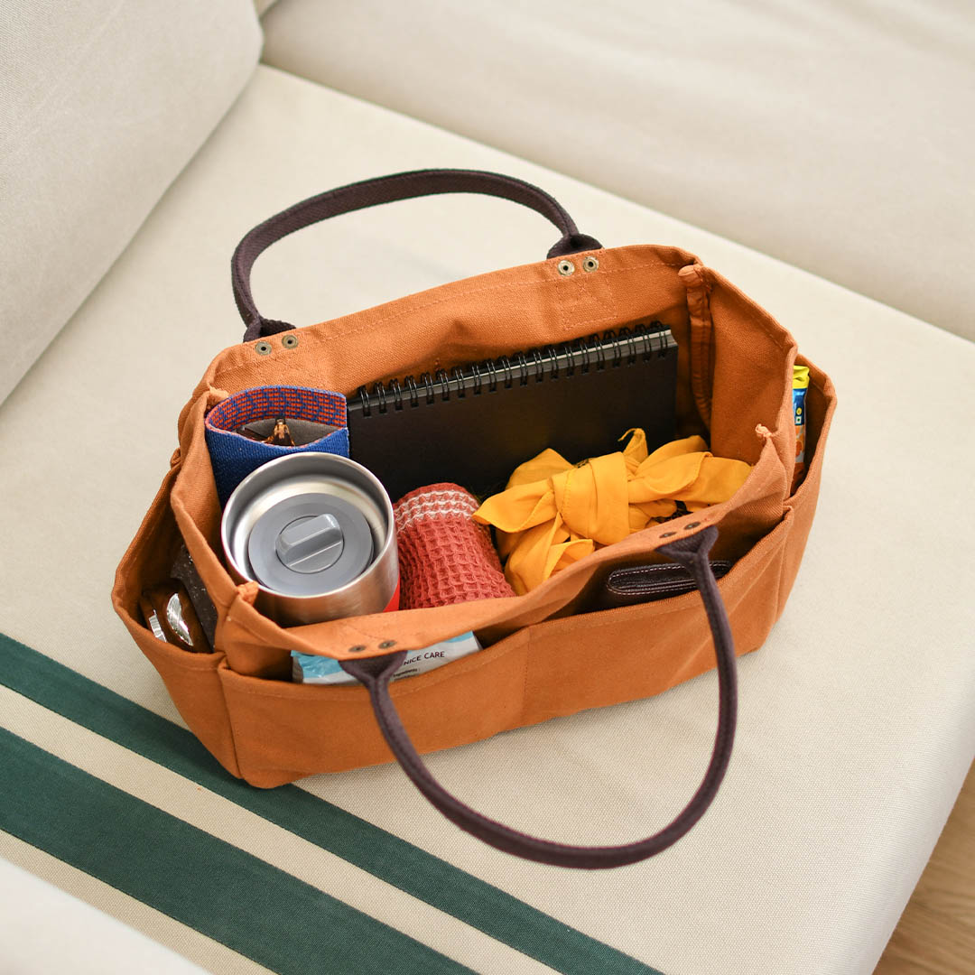 An orange fabric organizer bag with dark brown handles sits on a cream-colored sofa, holding various travel items like a notebook, a can, and a small red-and-white striped item.