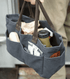 A dark gray canvas utility tote bag with brown handles, shown being carried by a person. The bag is filled with various items, including a beige bottle, a green travel mug, and small accessories, against a softly blurred outdoor background.