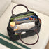 A black canvas organizer tote bag with dark brown handles, shown open and filled with various items including a ruler, notebook, pens, and a small bottle, resting on a light beige sofa.
