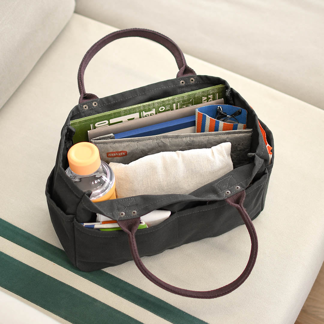 A black canvas organizer tote bag with dark brown handles, shown open and filled with various items including a ruler, notebook, pens, and a small bottle, resting on a light beige sofa.