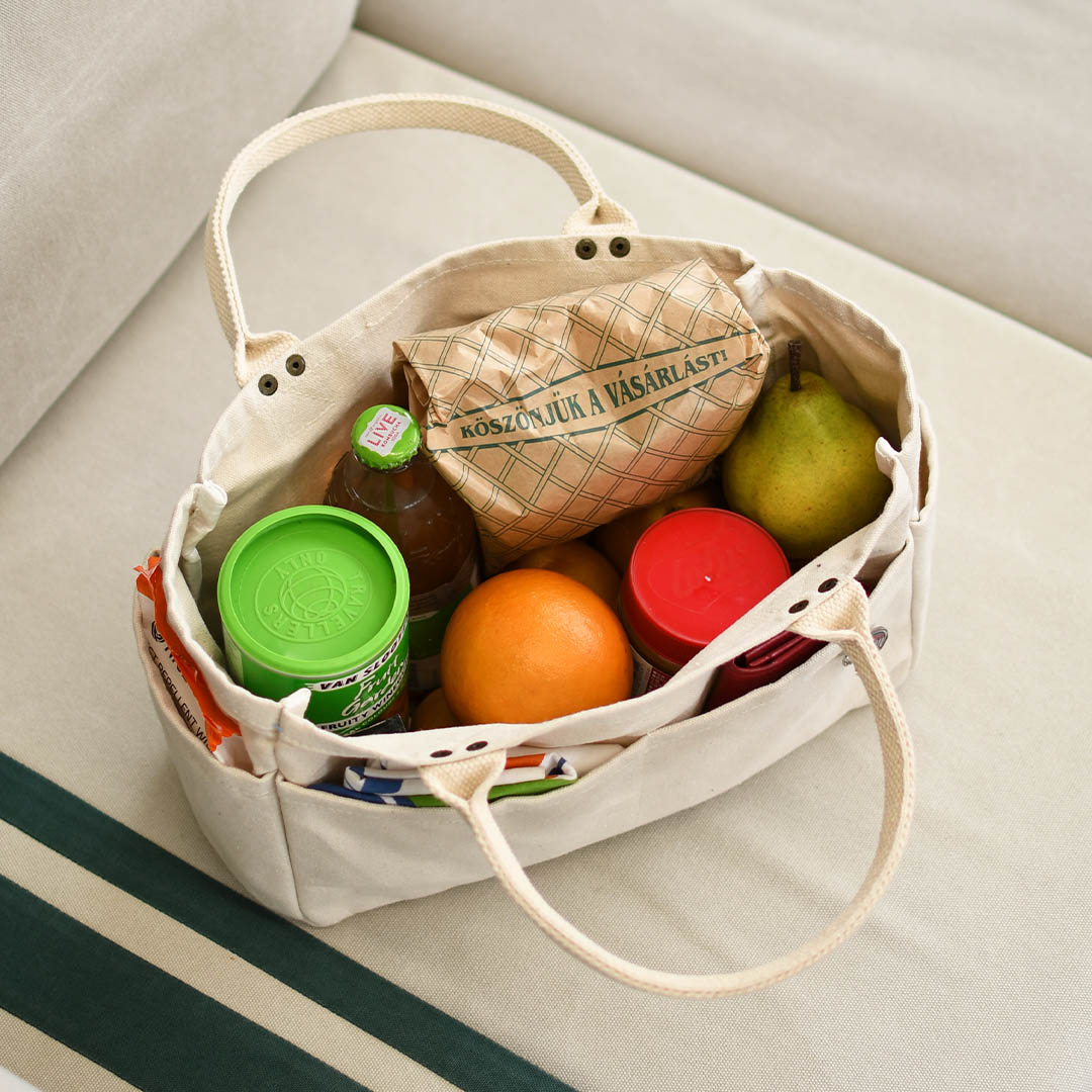 A cream canvas tote bag filled with groceries, including an orange, a green pear, a jar with a bright green lid, and a brown paper bag, placed on a light beige sofa.