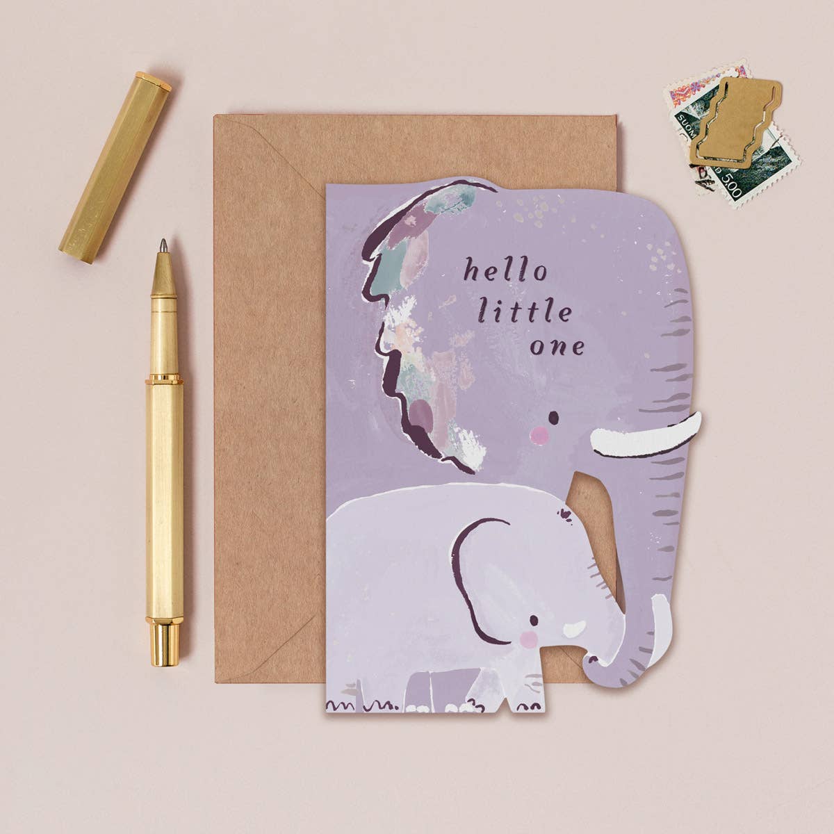 Greeting card with elephant design and 'hello little one' text, next to a pen and envelope on a beige background.