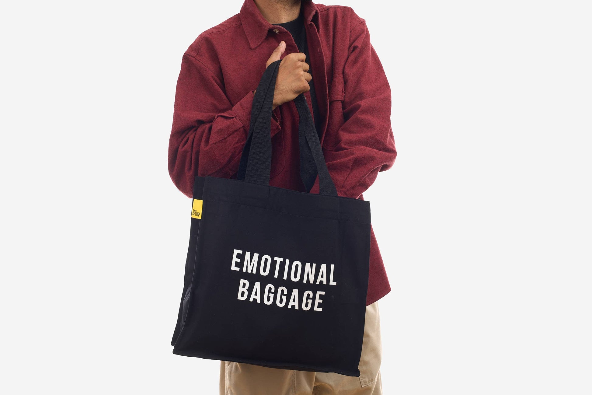 Person holding a black tote bag with 'EMOTIONAL BAGGAGE' text on a white background