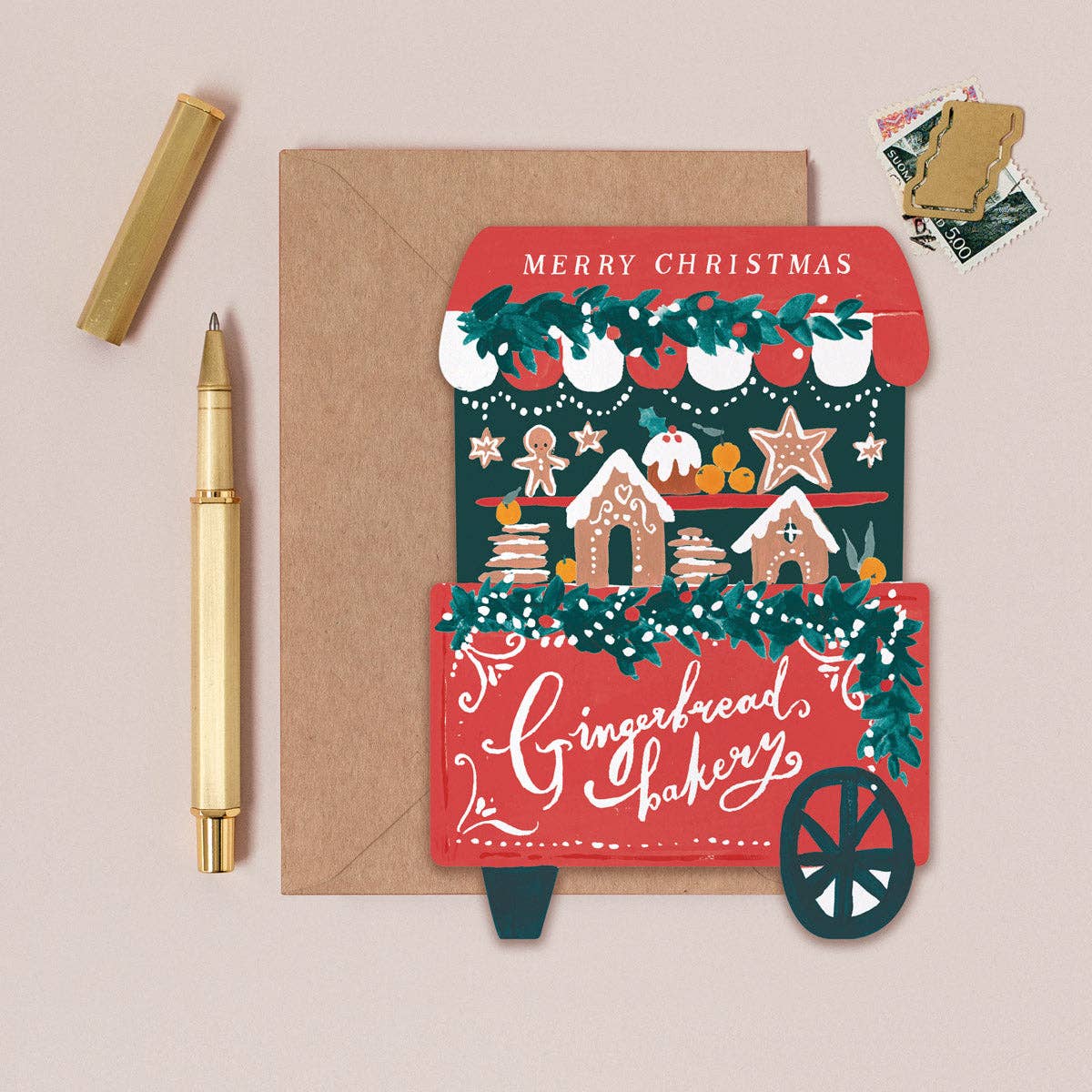 A Christmas-themed greeting card from 'The Gingerbread Bakery' styled in a flat-lay arrangement on a light beige background, accompanied by a brown envelope, a gold pen, and a gold cap.