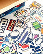 Collection of pixel art stickers on a white surface with a cardboard box in the background.