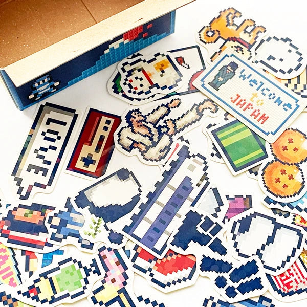 Collection of pixel art stickers on a white surface with a cardboard box in the background.