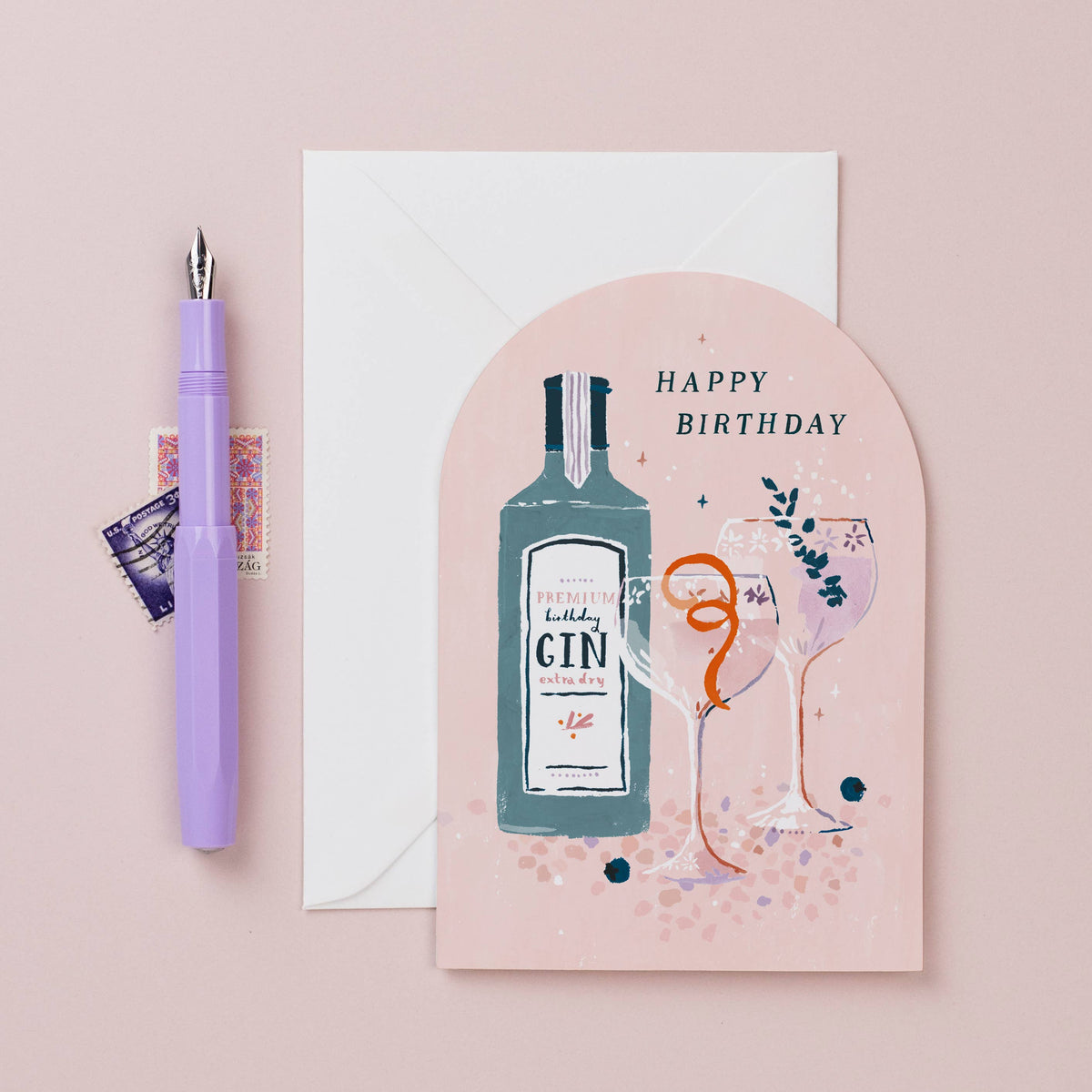 A pink arched birthday card with an illustration of a gin bottle, two champagne glasses, and the words 'HAPPY BIRTHDAY,' shown with a white envelope and a lavender fountain pen on a pale pink background.
