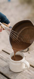 Person pouring chocolate mixture from a metal pot into a white mug on a wooden surface.