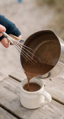 Person pouring chocolate mixture from a metal pot into a white mug on a wooden surface.