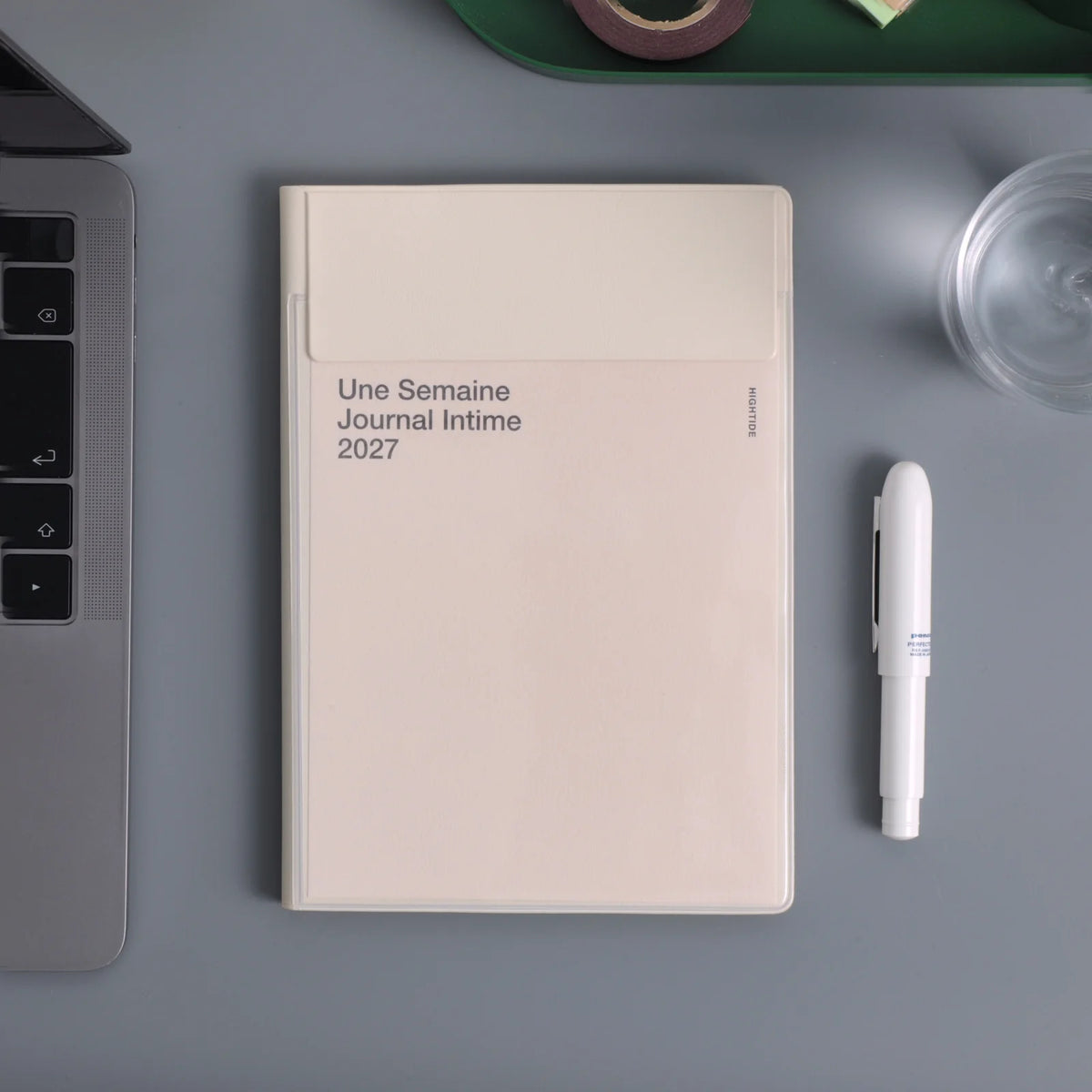 A cream-colored weekly journal with the French title 'Une Semaine Journal Intime 2027' lies on a gray desk, styled with a white pen, part of a laptop, and a glass cup.