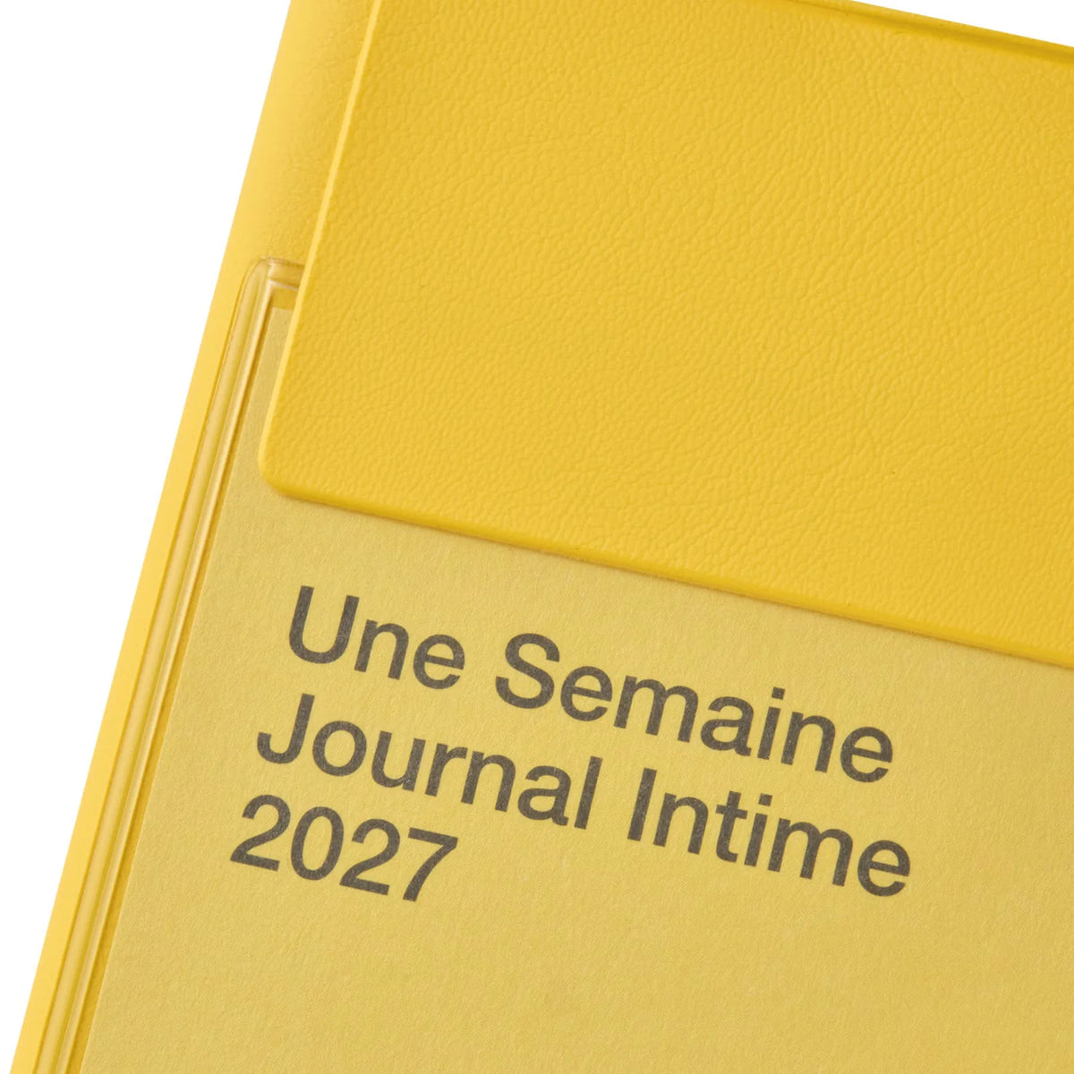 Close-up of a yellow textured journal cover with the French text 'Une Semaine Journal Intime 2027' printed in dark gray on a white background.