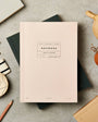 Pink notebook with branding on a gray surface with stationery items