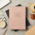 Pink notebook with 'Notebook' text on a gray surface with stationery items.