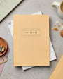 Beige notebook on a gray surface with office supplies and a cup of coffee.