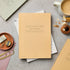 Beige notebook on a gray surface with office supplies and a cup of coffee.