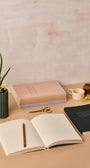 Open notebook with a pen, closed books, and decorative items on a beige surface.