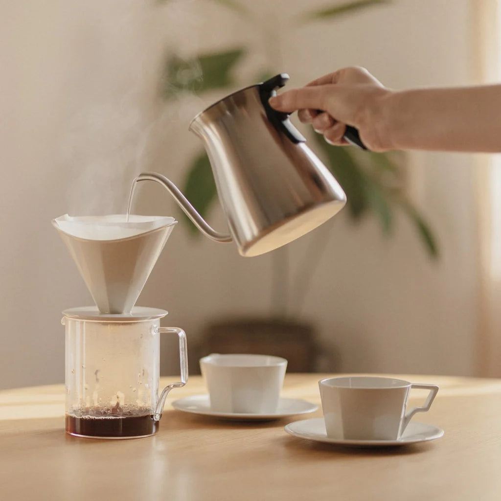 Person pouring coffee from a stainless steel kettle into a glass carafe on a wooden table.