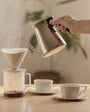 Person pouring coffee from a stainless steel kettle into a glass carafe on a wooden table.