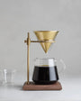 Coffee brewing setup with glass carafe, brass dripper, and wooden stand on a gray background