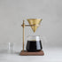 Coffee brewing setup with glass carafe, brass dripper, and wooden stand on a gray background