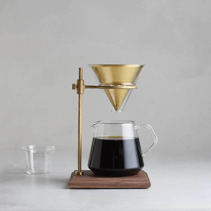 Coffee brewing setup with glass carafe, brass dripper, and wooden stand on a gray background