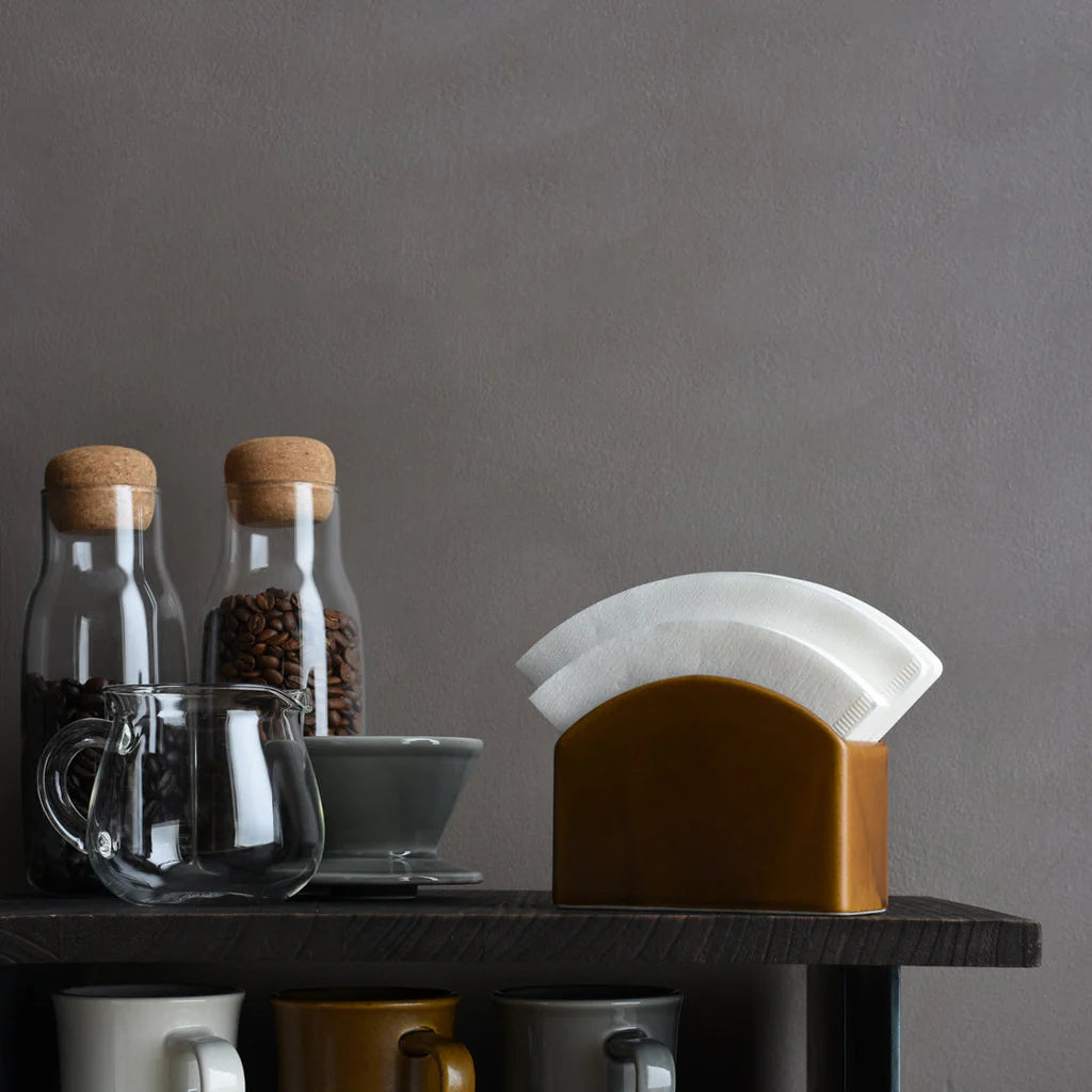 Coffee-making setup with glass jars, coffee beans, a filter, and mugs on a dark surface against a gray background.