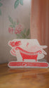 Decorative card with a red and white design of a woman in a bathtub on a wooden surface.