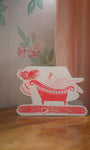 Decorative card with a red and white design of a woman in a bathtub on a wooden surface.