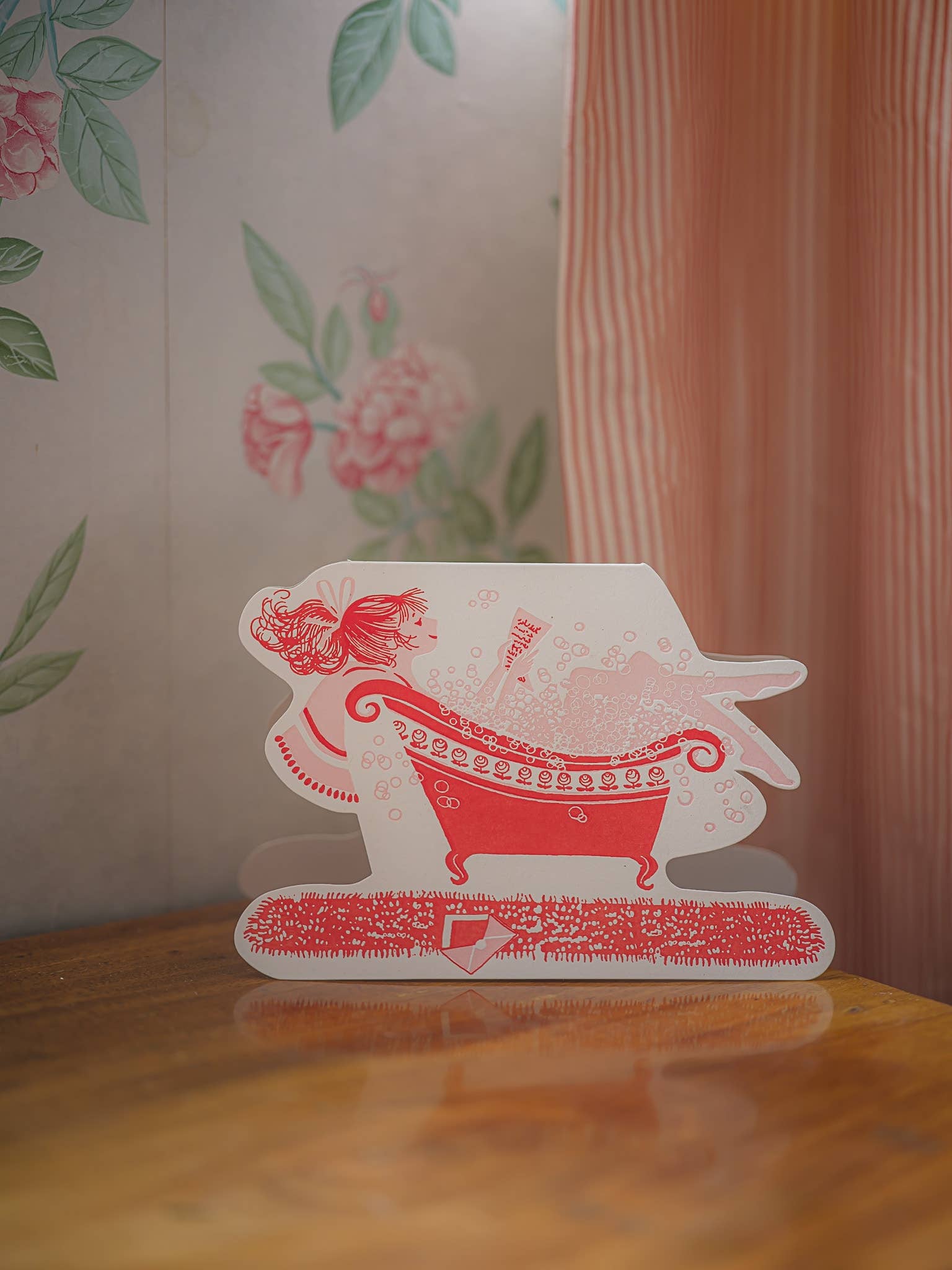 Decorative card with a red and white design of a woman in a bathtub on a wooden surface.