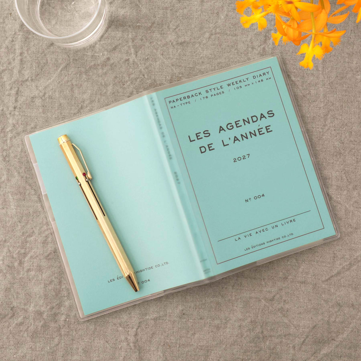 A clear plastic sleeve holding a light blue yearly planner and a gold pen, photographed on a textured gray fabric surface with a glass and yellow flowers as props.