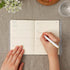 An open planner or journal with cream pages lies on a gray fabric surface, with a hand holding a white pen writing in the right page. A second hand rests on the left page.