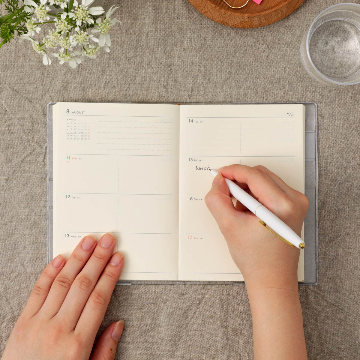 An open planner or journal with cream pages lies on a gray fabric surface, with a hand holding a white pen writing in the right page. A second hand rests on the left page.