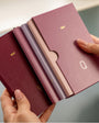 Four slim 2027 pocket calendars in burgundy, mauve, dusty pink, and lavender, held in two hands to show the thickness and texture of the covers.