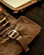 A partially unrolled tool roll with a brown waxed canvas exterior, aluminum hook fastener, and multiple pockets containing pens and other tools.